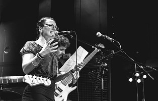 Photo : une femme, adulte mais jeune, parle avec présence au micro. Cheveux attachés, lunettes, chemisier, jupe : ses mains sont levés près du micro et tiennent aussi une feuille.  Derrière un musicien à la basse électrique. On voit aussi le bout d'une guitare électrique.