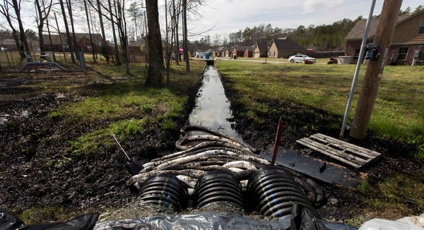 Photo d'une sortie de trois tuyaux où visiblement du pétrole a coulé dans un petit ruisseau. Le gazon est imbibé de pétrole. Forêt et maisons à côté.