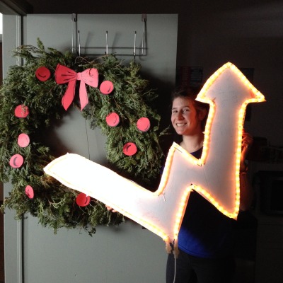 Photo : grande couronne de Noël [cercle de branches de cèdre. Une jeune femme tient une grosse flèche lumineuse indiquant une hausse: flèche qui rappelle le symbole d'Hydro-Québec]