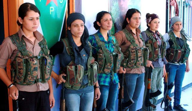 Six femmes sont en ligne, tendant à leur côté une AK47; cheveux longs, jeunes, jolies. Vêtements civils, blue jeans, mais harnais de combat et munitions. Le symbole de la communauté Kobané (kurde) derrière.