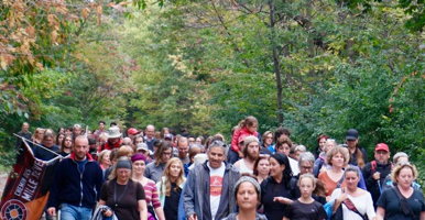 Photo : une centaine de personnes, marchant ensemble, vues de face dans une forêt boréale au soleil. Plusieurs femmes, âges variés. On peut discerner M. Vollant au milieu vers l'avant discutant avec une jeune femme.
