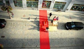 Photo officielle de l'événement : une rue vue du ciel, environ 50 pieds au-dessus de six personnes qui ont installé un espace sur le stationnement (une place) devant un magasin et sur le trottoir. Une voiture noire de chaque côté. Une large ligne rouge traverse la zone installée.