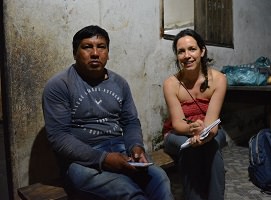 Photo d'un homme d'air musclé à la peau brune foncée, cheveux noirs. Marie-Josée à côté, cheveux longs bruns tressés, souriante, peau pâle. La maison a des murs de terre blancs usés par le temps et des fenêtres en vieux bois, le tout donne une texture intéressante.