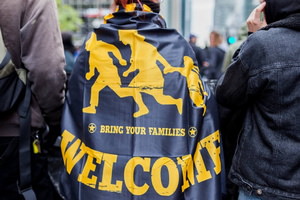 Photo : une june femme porte une cape avec le dessin jaune d'enfants qui courent. « Bring your families ». Deux jeunes hommes avec elle : ce semble être sur la rue lors d'une manifestation.