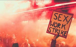 Photo : dans une manif ou une émeute, couverte de fumée, un discerne une personne qui tient une pancarte bien haute se lisant « Sex / Work Strike ».