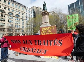 Photo d'un rassemblement devant un monument à Montréal : deux femmes tiennent une bannière rouge se lisant « Non aux traités coloniaux ».