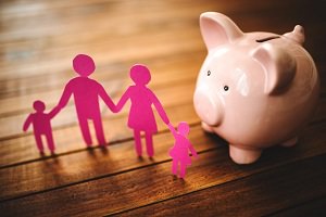 Photo : banque en forme de petit cochon rose sur une table en bois brun. Devant se tient la forme d'une petite famille humaine, découpée de papier rose.