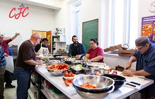Photo de sept personnes dans la cuisine, autour d'une table avec des bols en métal et divers fruits et légumes. Logo CJC. Âges variés, etc.