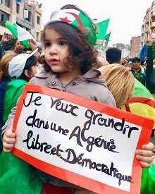 Photo d'une jeune enfant algérienne tenant une affiche : « Je veux grandir dans une Algérie libre et démocratique ». Elle a les cheveux bruns, un peu frisés aux bouts, et a un bandeau aux couleurs du pays. Une foule derrière elle (manif).