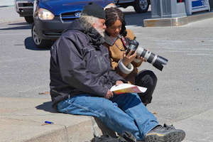 Photo : deux personnes assieds sur un trottoir près de voitures. Une jeune femme, peau brune, tenant une caméra et la montrant à un monsieur aux cheveux et barbes blanches et tenant des feuilles de papier.