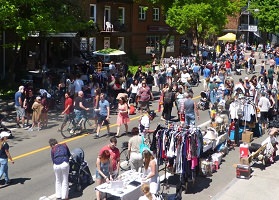 Photo : foule de gens sur la 3e Avenue ; des kiosques ici et là ; il fait beau soleil ; arbres verts. 