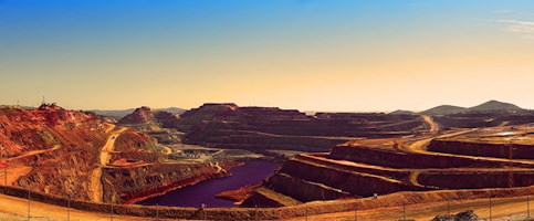 Photo panoramique : vaste mine à ciel ouvert, de sable orangé, en étages. Une rivière ou lac mauve au bas.