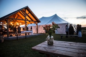 Photo : sur du gazon, à l'extérieur le soir, un gazebo (pavillon) en bois et illuminé orange, une table à pique-nique avec bouquet de fleurs, femmes qui discutent en cercle, autre gazebo (tente) blanche.