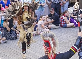 Photo : une femme souriante et un homme de dos, vêtus de cuir beige et de plumes, dansent ensemble. Autour, une foule d'enfants et parents.