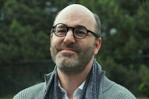 Photo de M. Deneault devant des arbres verts. Lunettes noires rondes, barbe légère, air souriant, presque chauve.