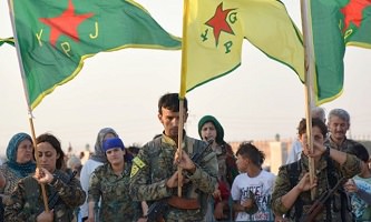 Des femmes et hommes militaires kurdes, tenant des drapeaux du YPJ et YGP, baissent la tête pour honorer les défunts. Une foule civile derrière.