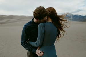 Photo : jeune homme, femme aux cheveux longs brun, visages un sur l'autre, sur un désert au sable fin beige et montagnes au loin.