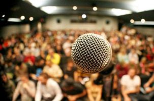 Photo d'un micro devant une salle comble. Le focus est sur le micro et la foule assise derrière est floue.