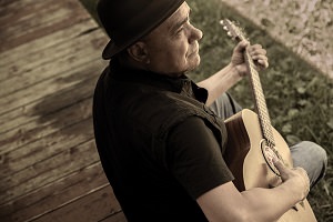 Photo de Florent Vollant, assied sur un balcon en bois, jouant une guitare. T-shirt et veste noir, chapeau brun style un peu rond ou melon.