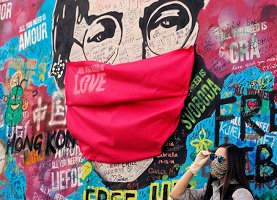 Photo d'arts et graffitis sur un mur extérieur : un masque rouge géant sur un visage géant portant des lunettes avec le mot « Love », « Hong Kong », etc. Une jeune femme à côté pose avec un masque et des lunettes de soleil.