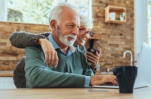Photo d'un couple d'environ 60 ans regardant ensemble un ordinateur portable, dans une belle cuisine ensoleillée. Un monsieur à la barbe blanche et, derrière lui, une dame souriante tenant une tasse.