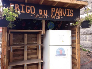 Photo 2019 du frigo en question, placé dans un beau cabanon en bois, avec deux pots de fleurs, et une panneau « Frigo du parvis Saint-Roch ».