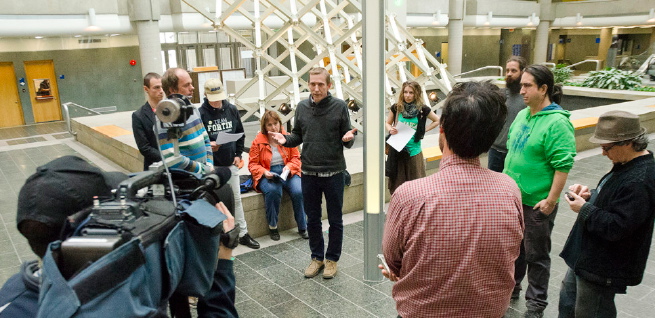 Photo du point de presse quand Sébastien Harvey, de la Ligue des droits et libertés, s'adresse aux médias. Un caméraman est vue de dos. Devant, il y a 8 militant-es et journalistes indépendants. André Bérubé, le plaignant, écoute M. Harvey.