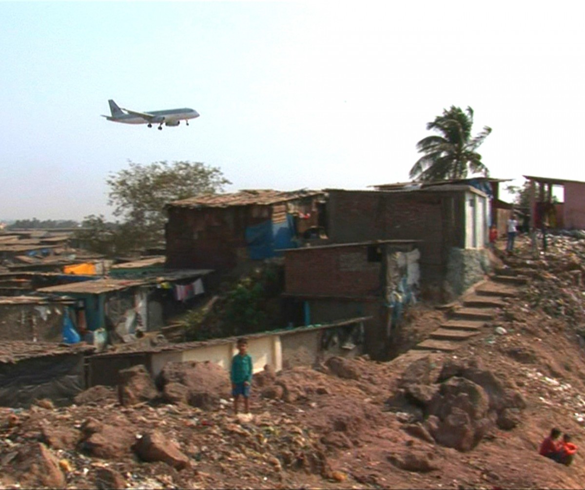 Un avion décollant survole un bidonville quelque part en Inde, un enfant se tient debout à l'horizon.