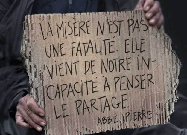 Deux mains tiennent un vieux carton avec les mots : La misère n'est pas une fatalité. Elle vient de notre incapacité à penser le partage. - Abbé Pierre.