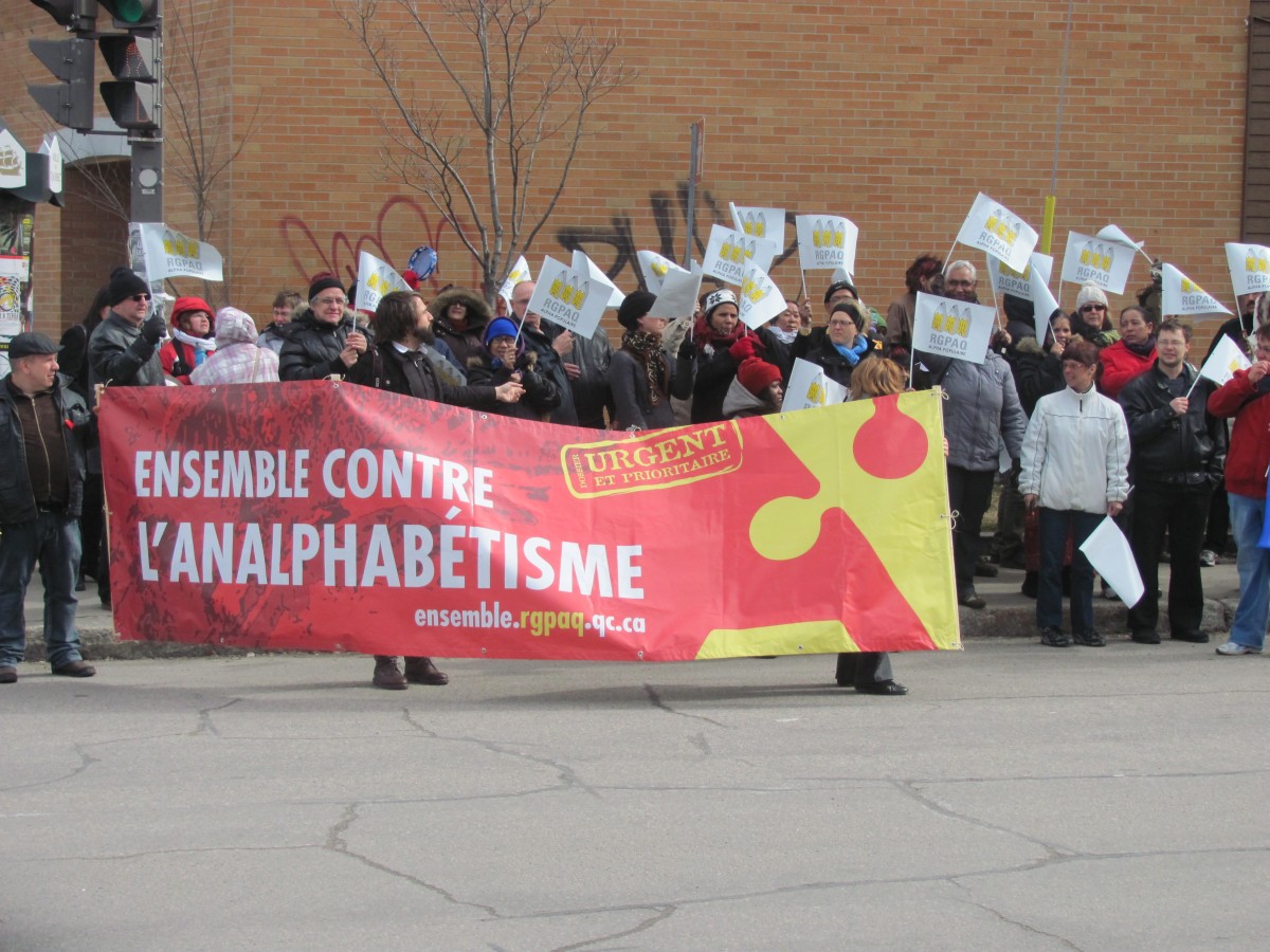 Photo : une foule tenant des fanions du RGPAQ et une bannière « Ensemble contre l'analphabétisme - Urgent et prioritaire ».