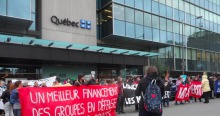 Photo : devant le ministère, sur le Québec on voit le drapeau du Québec, une foule de gens se tiennent en ligne unie avec des bannières, dont la plus lisible dit: Un meilleur financement des groupes en défense collective des droits.