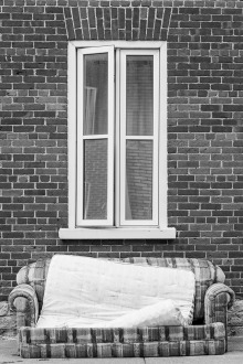 Photo : fênetre sur un mur de brique ; un matelas sur un divan est à l'extérieur, devant la fenêtre. Noir et blanc.