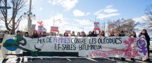 Photo : longue banderole devant une manif de femmes portant le nom de l'événement. Il faut beau soleil. La banderole a deux oeuvres artistiques : une plante animée entoure la Terre pour la protéger d'un oléoduc noir. Il y a des symboles incompréhensibles rose-rouge, de style un peu autochtone, avec les mots Libérons nos corps, notre Terre et nos territoires.