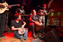 Photo : cinq personnes sur une petite scène. Éclairage rouge. Un jeune homme à la peau pâle joue du tam tam. Une dame plus âgée à la peau foncée joue aussi du tam tam avec lui. Un chanteur les accompagne, un bassiste et un guitariste, tous à la peau foncée d'origine africaine.