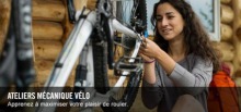 Photo : une jeune femme aux cheveux long brun-roux travaille sur un vélo argenté. Maison en bois ronds derrière.