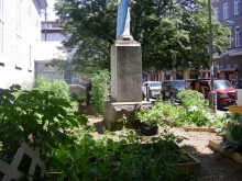 Photo du potager collectif, autour d'une statue de la Vierge-Marie au centre, vue de profil. On voit les voitures stationnées le long de la rue.