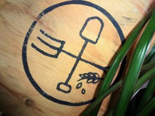 Logo de Verdir Saint-Roch étampé sur bois : un cercle noir avec une fourche et une pelle croisées. Un brin de blé dessous.